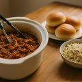 Shredded pulled pork in a slow cooker insert with buns and coleslaw nearby