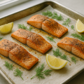 Four baked salmon fillets on a parchment-lined sheet pan with lemon wedges and dill